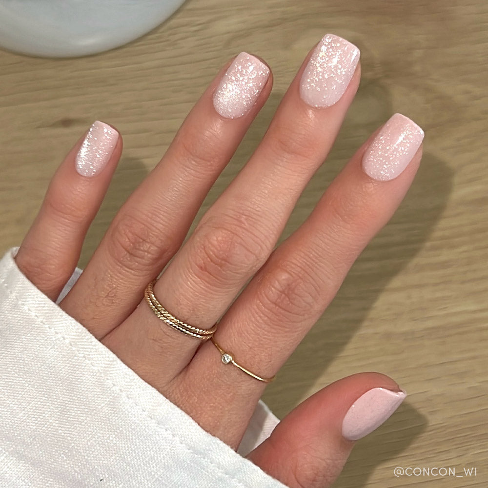 A hand with short squoval nails wears glamnetics Pinky Promise—a pale pink, shimmering glitter manicure with a blush velvet finish. Two gold rings and a white long-sleeve shirt rest against a light wooden background.